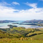 Panoramic view of Whakaraupō / Lyttelton Harbour from the surrounding hills, showing green rolling farmland, patches of forest, and the calm harbour waters under a partly cloudy sky.