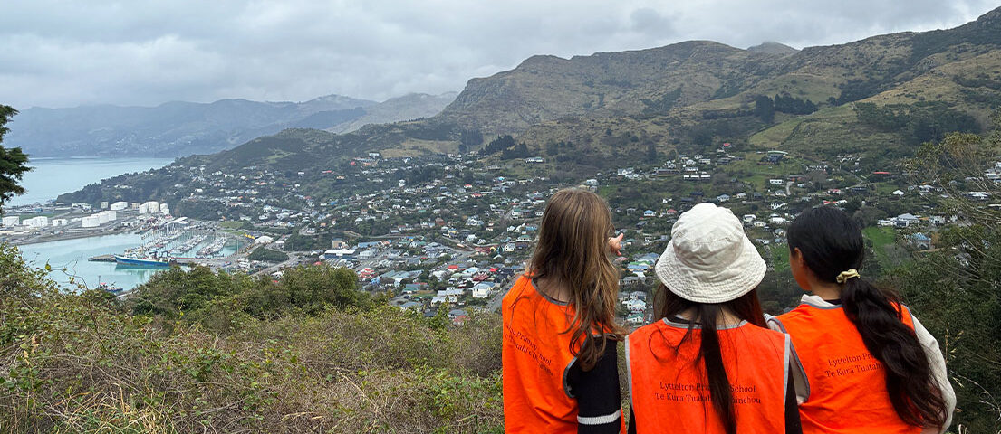 Three students in orange safety vests look out over Lyttelton and Whakaraupō Lyttelton Harbour from Port Saddle.
