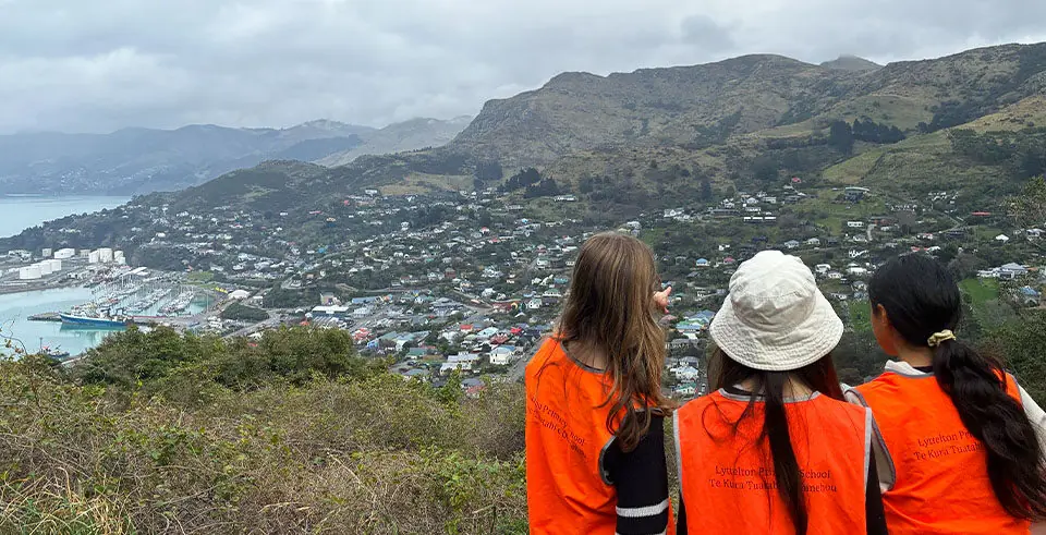 Three students in orange safety vests look out over Lyttelton and Whakaraupō Lyttelton Harbour from Port Saddle.